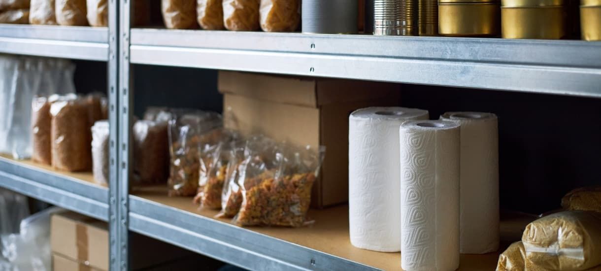 Stainless steel commercial kitchen shelving stocked with paper towel rolls, cardboard storage boxes, sealed bags of dry goods, and canned foods in a clean restaurant dry storage pantry. Metal shelving unit stocked with boxed and bagged food supplies in restaurant pantry.